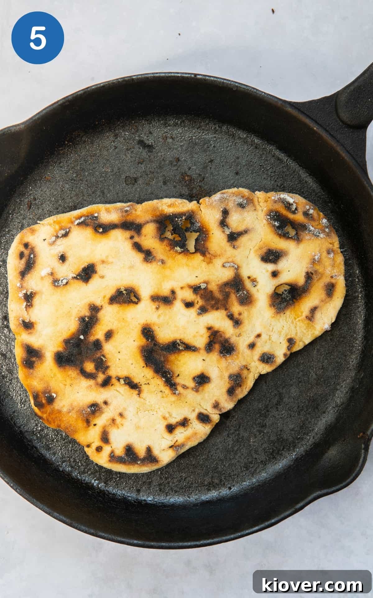 A gluten-free naan bread cooking in a large black frying pan on high heat, starting to bubble and char slightly.