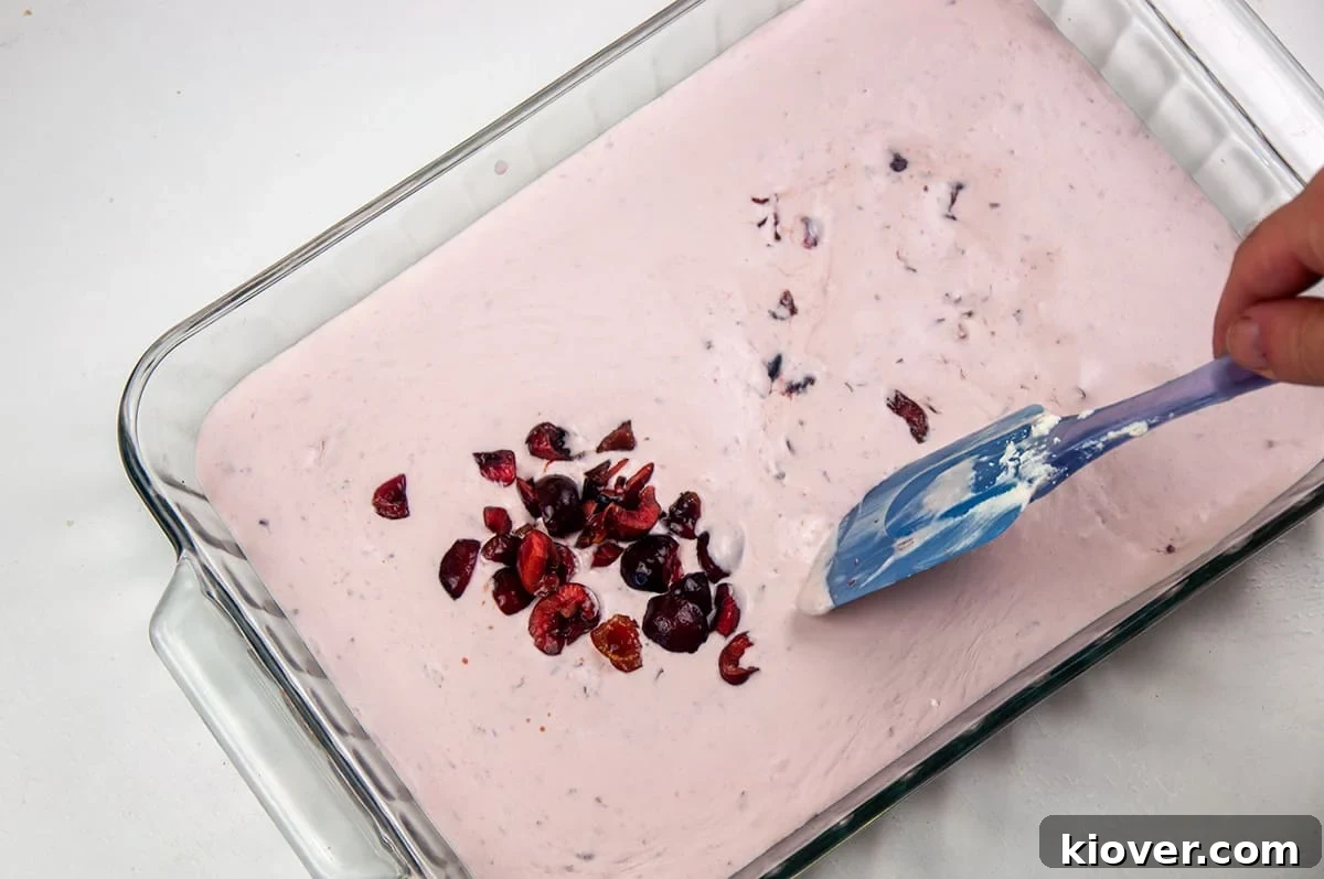 A 9x13 baking dish filled with unfrozen ice cream and a blue spatula mixing in chopped cherries.