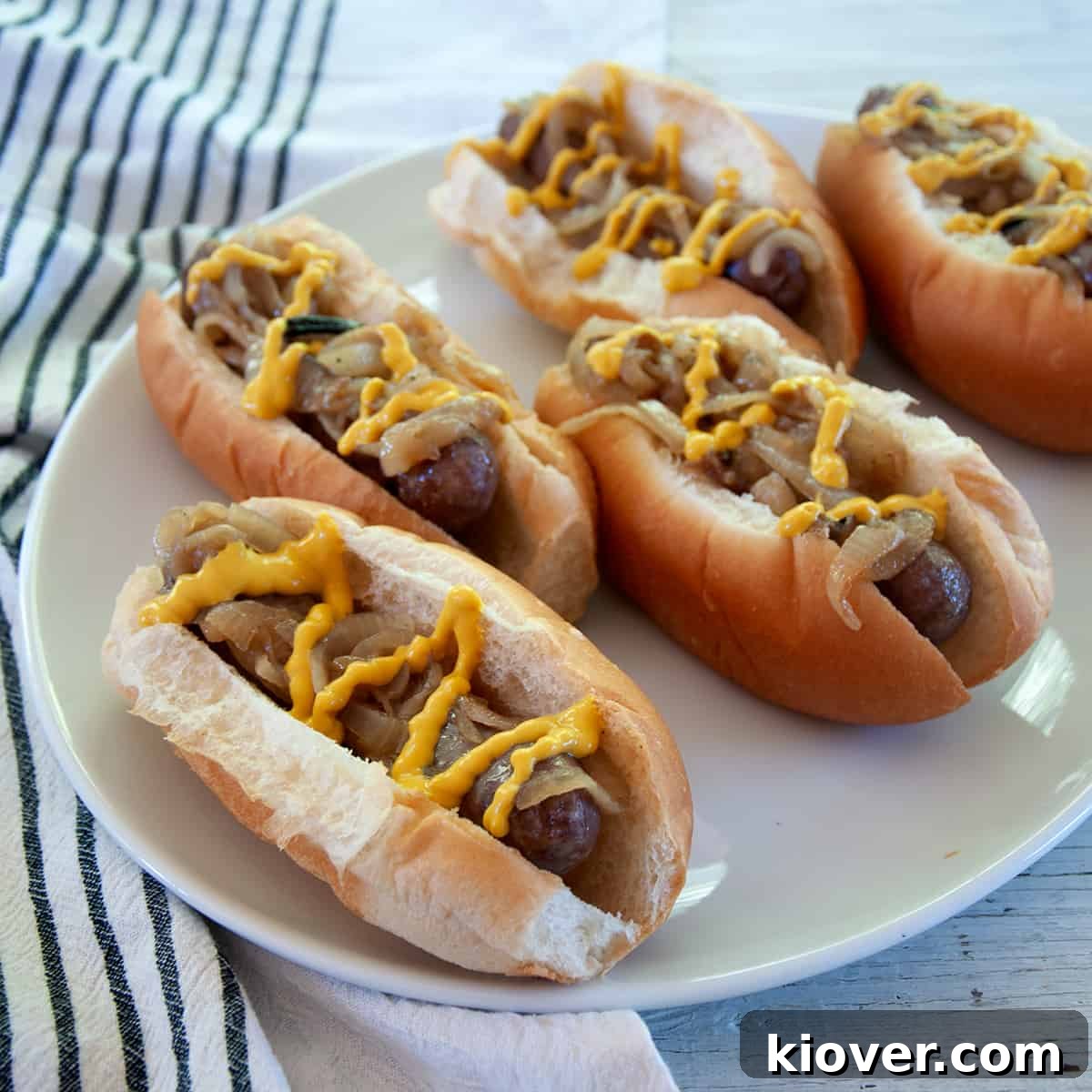 Five perfectly grilled bratwursts nestled in toasted buns, generously topped with sweet caramelized onions and a drizzle of classic yellow mustard.