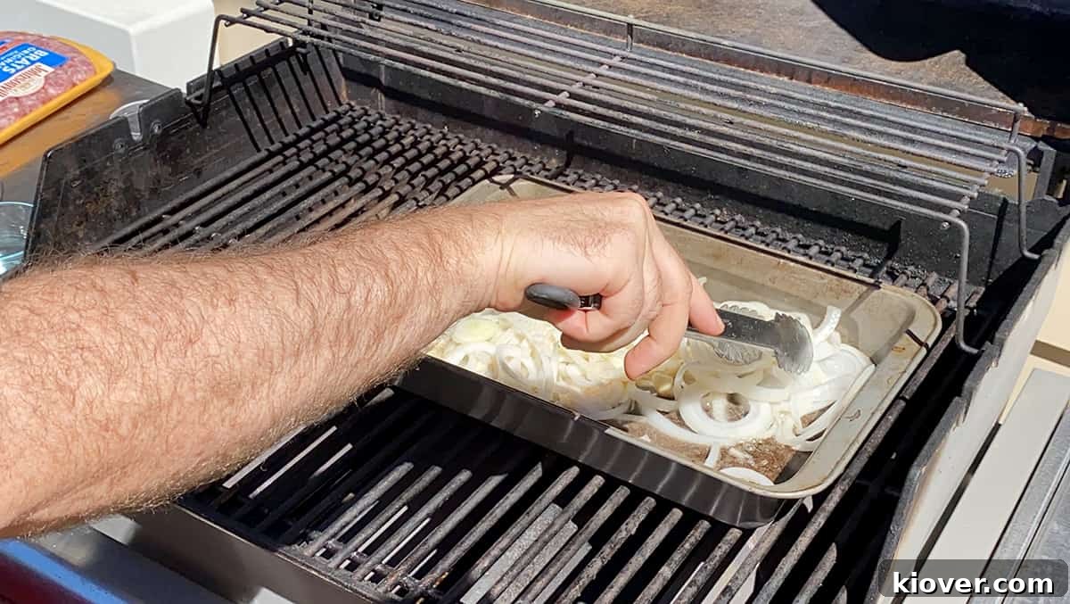 A pair of tongs expertly mixing thinly sliced white onions in a baking dish filled with melted butter and minced garlic on a grill top.