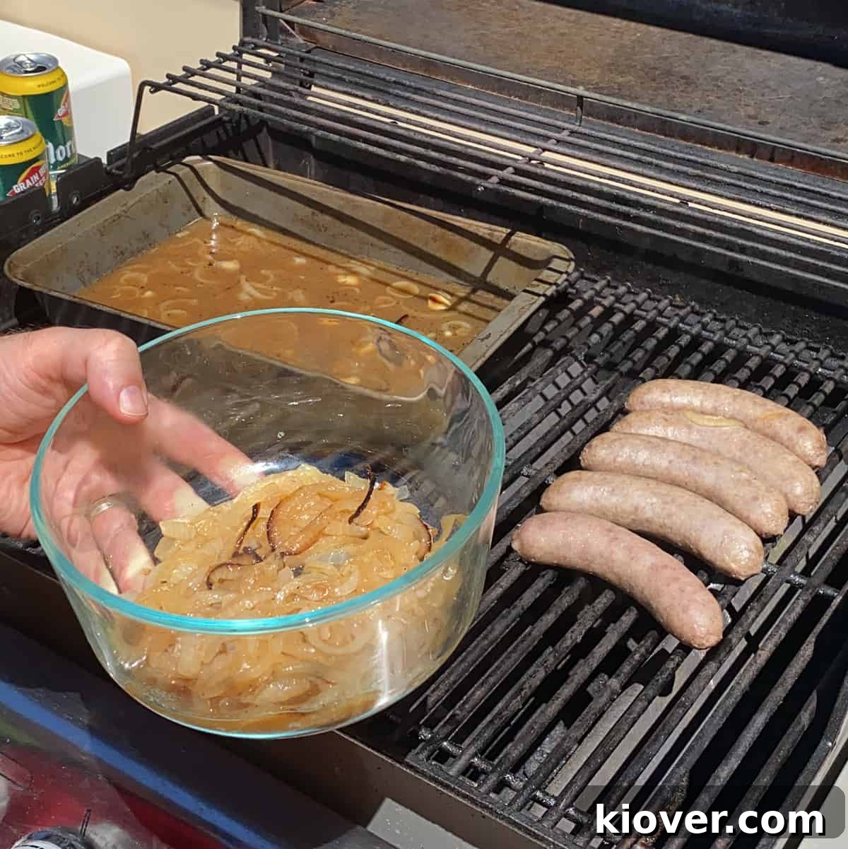 Five sizzling brats cooking on the grill grates alongside a bowl of golden, caramelized onions that have been removed from the beer bath.