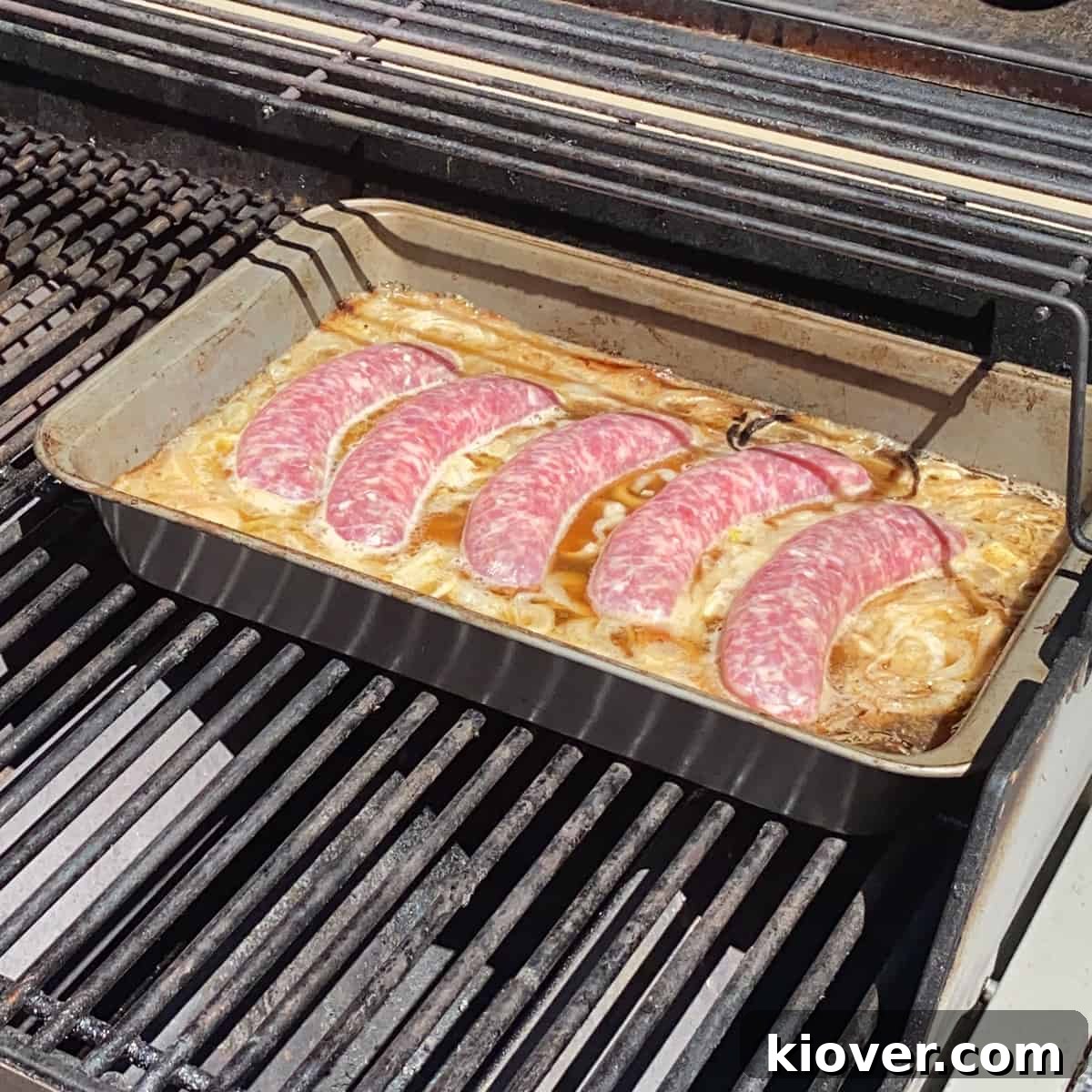 Five plump, uncooked bratwurst sausages gently simmering in a pan filled with beer, caramelized onions, and garlic.