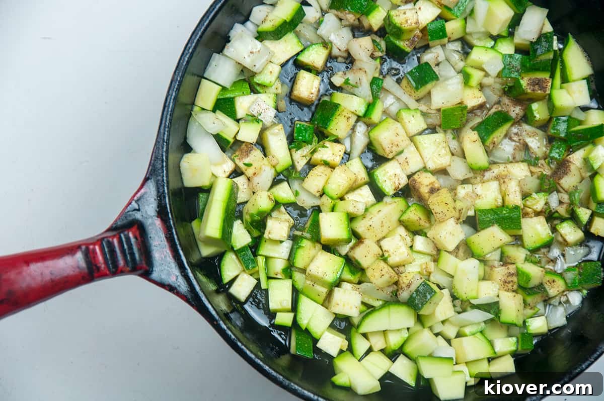 A cast iron skillet holds frying zucchini, onions, and aromatic herbs, gently cooking to develop their flavors.