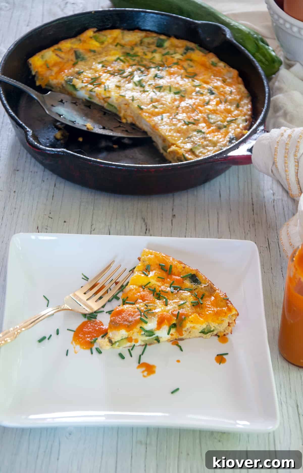 A single slice of Zucchini Frittata, elegantly topped with a drizzle of hot sauce, rests on a plate while the remainder of the frittata fills the cast iron pan in the background.