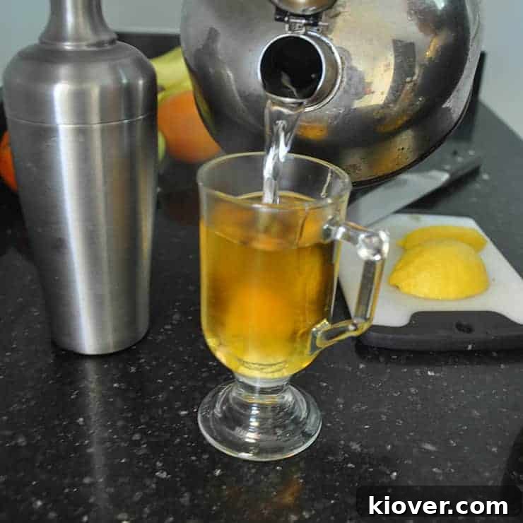 Pouring hot water into hot toddy on black countertop with shaker and lemon slices on the side