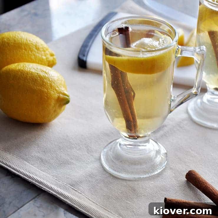 Backlit image of a steaming Hot Toddy on a linen tablecloth, accompanied by two fresh lemons and fragrant cinnamon sticks.