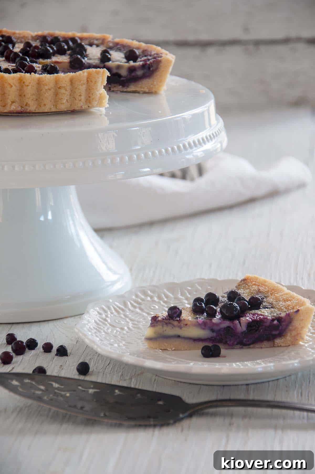Slice Blueberry Tart Single slice of blueberry tart on a white plate with the rest of the tart on a platter.