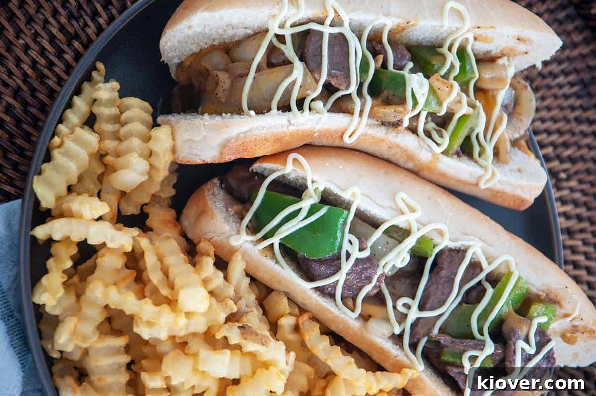 Two perfectly assembled Bison Philly Cheesesteaks on a white plate, accompanied by a generous serving of crispy french fries.