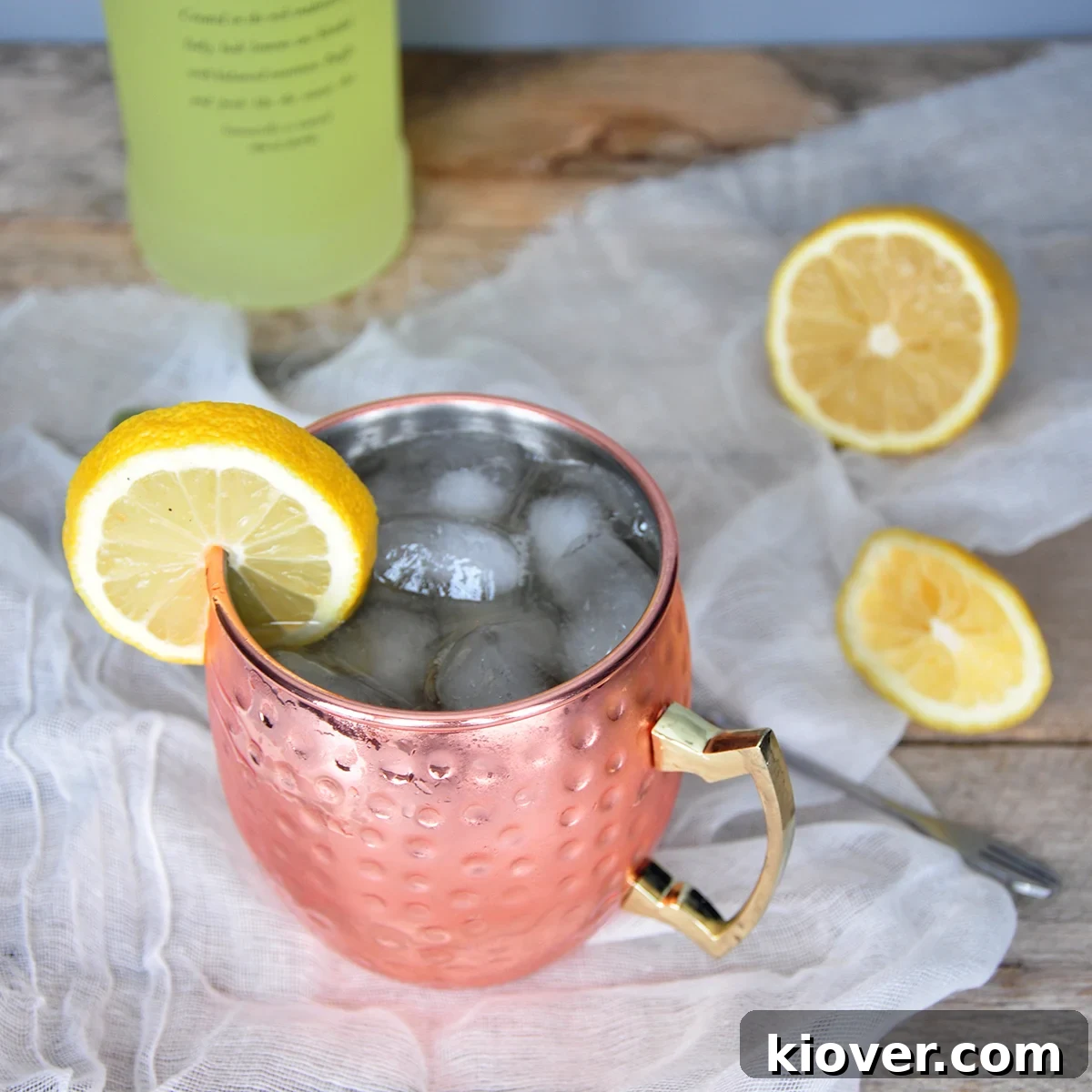 A refreshing Limoncello and Lavender Moscow Mule cocktail in a copper mug, garnished with a lemon slice and lavender sprig, on a rustic wooden table with a bottle of limoncello in the background. A stirring spoon and fresh lemon slices complete the scene.