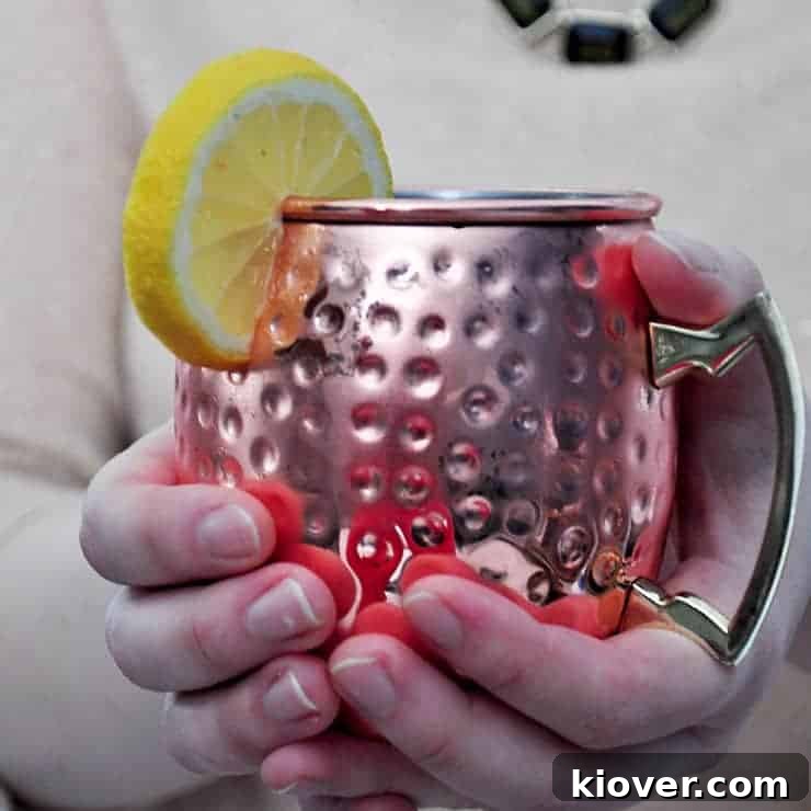 A person's hands holding a shimmering copper mug filled with a Limoncello and Lavender Moscow Mule, garnished with a fresh lemon slice, against a blurred background.