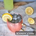Limoncello and Lavender Moscow Mule on a wood table with a bottle of limoncello in the background. There is a sliced lemon on the table and a stirring spoon.