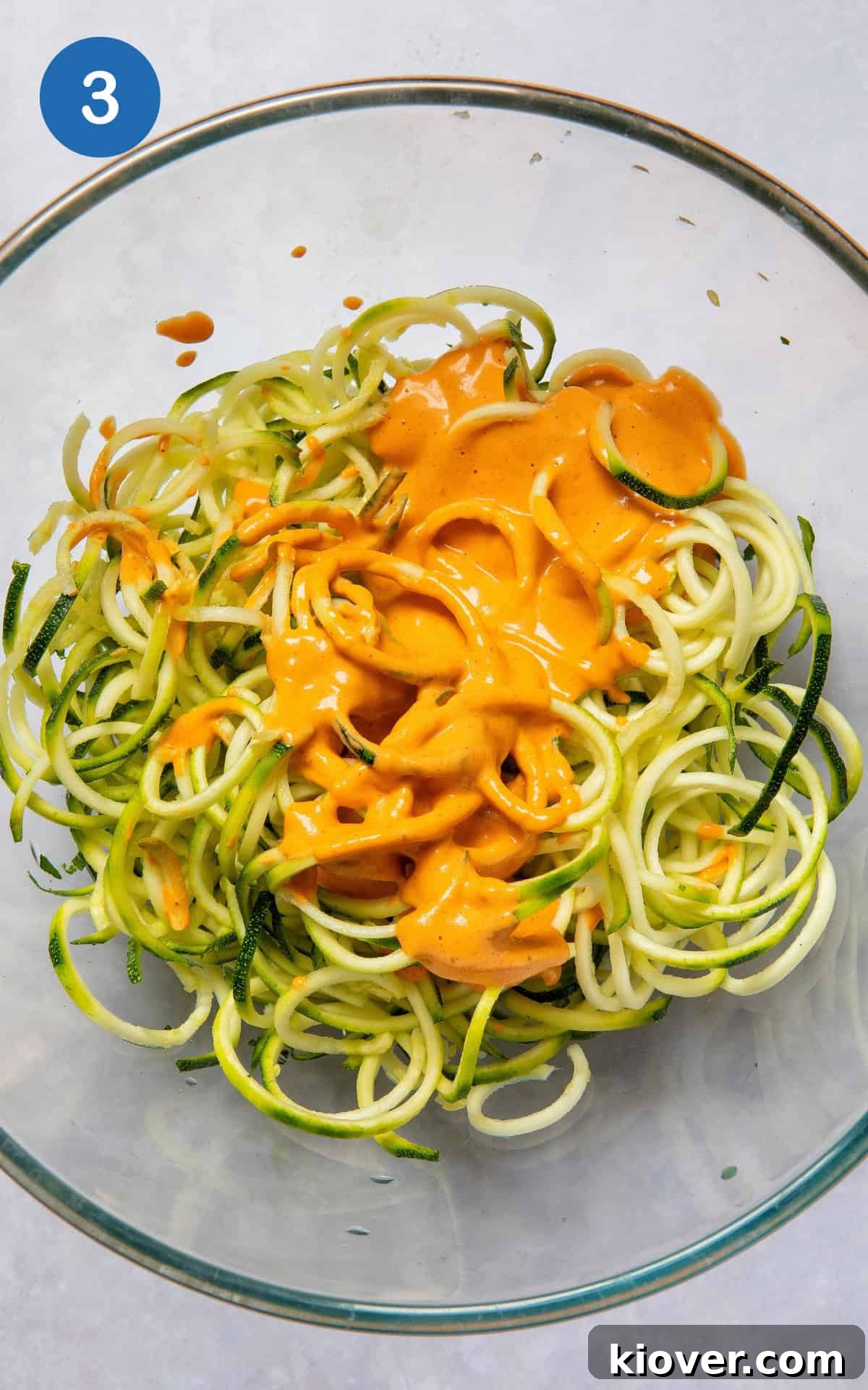 The creamy cashew sauce being poured generously over the spiralized courgetti and other salad ingredients in a mixing bowl.