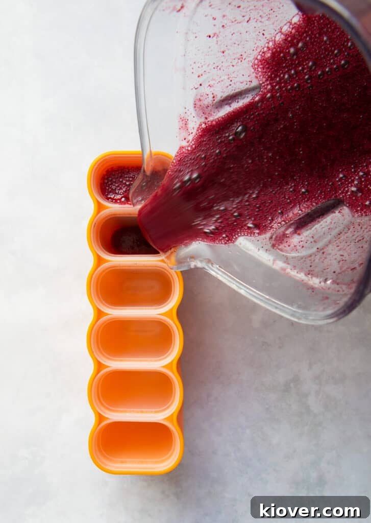 Vibrant blueberry juice being poured from a blender into multiple ice lolly molds.