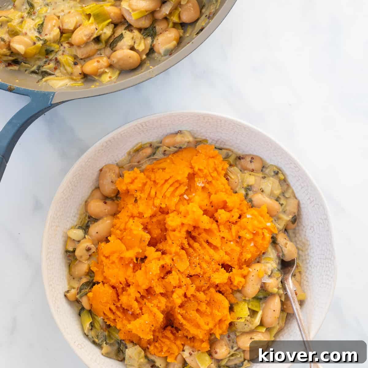 Closeup of a delicious Gluten-Free and Vegan Butterbean and Leek Pie topped with a creamy sweet potato mash, served in a white bowl with a silver spoon on a marble countertop.