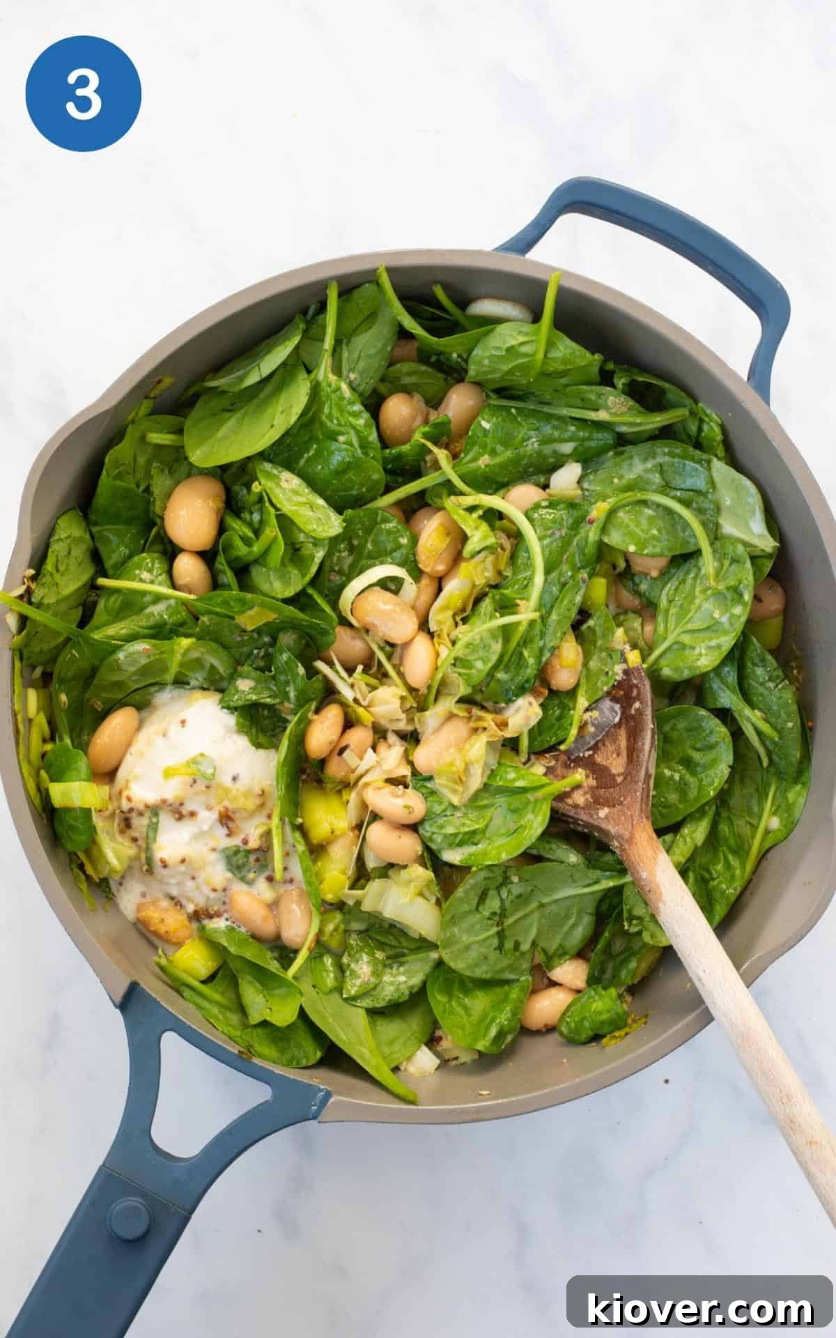 A mix of butterbeans, fresh spinach, coconut milk, and other pie ingredients simmering in a frying pan, stirred with a wooden spoon.
