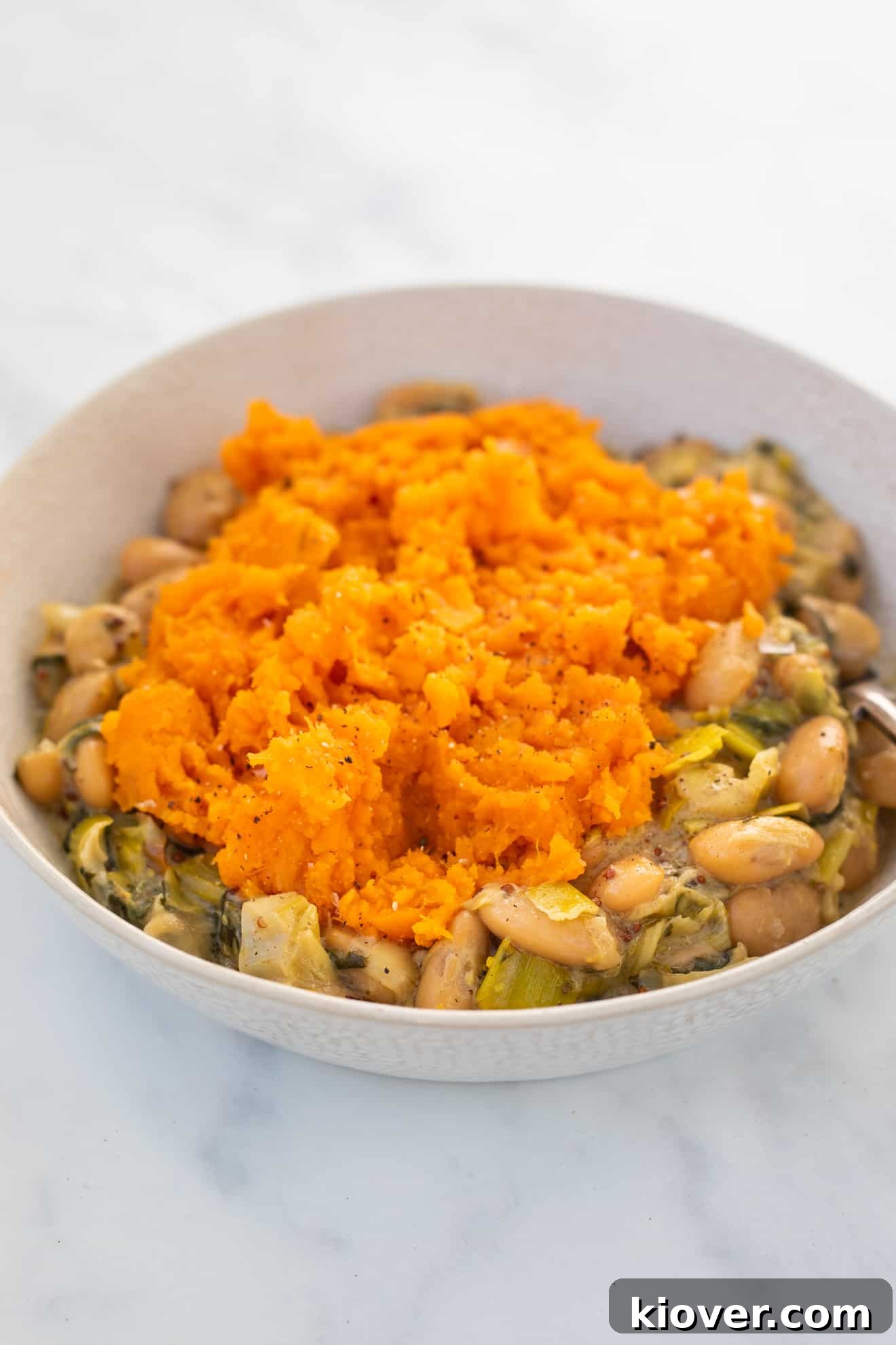 Side view of the finished Gluten-Free and Vegan Butterbean and Leek Pie, topped with a smooth sweet potato mash, presented in a white bowl with a silver spoon on a marble countertop, ready to be enjoyed.