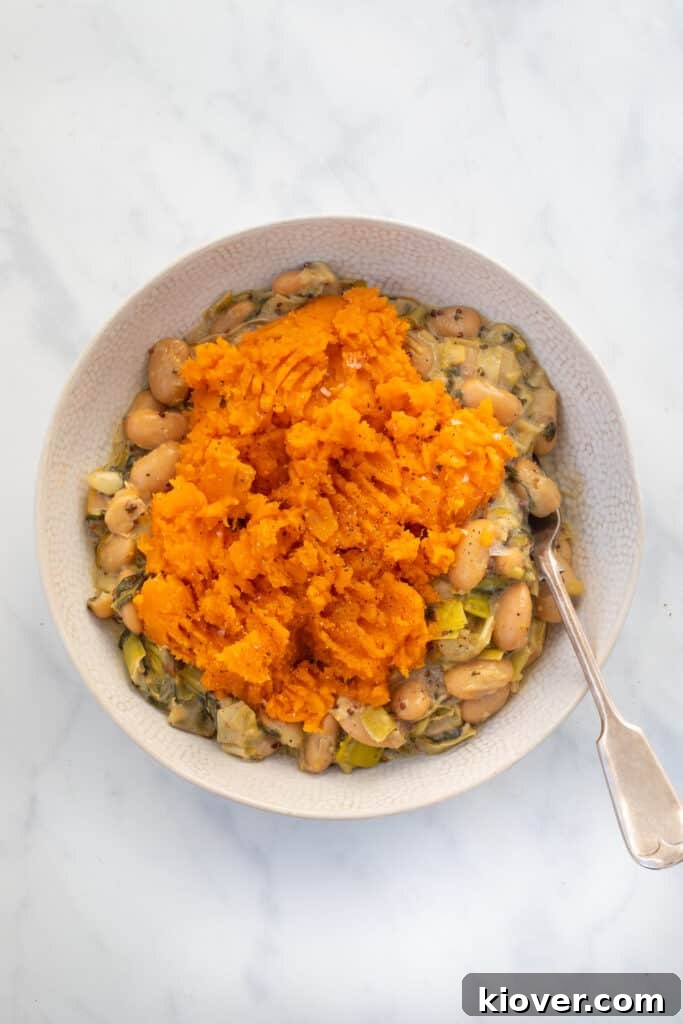 Overhead view of a delicious Gluten-Free and Vegan Butterbean and Leek Pie, generously topped with sweet potato mash, served in a white bowl with a silver spoon on a marble countertop. The pie is beautifully textured and inviting.