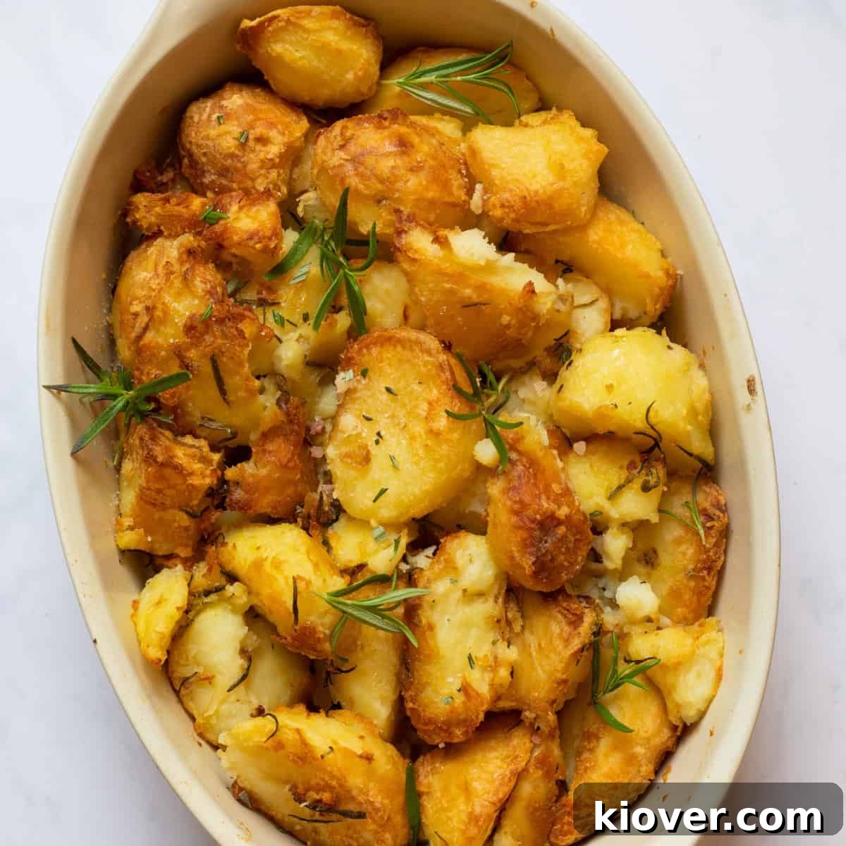 Crispy Rosemary Roasted Potatoes 2 Golden, crispy rosemary infused roast potatoes in a rustic baking dish on a white marble background, garnished with fresh rosemary sprigs, perfect for Christmas or a Sunday roast.
