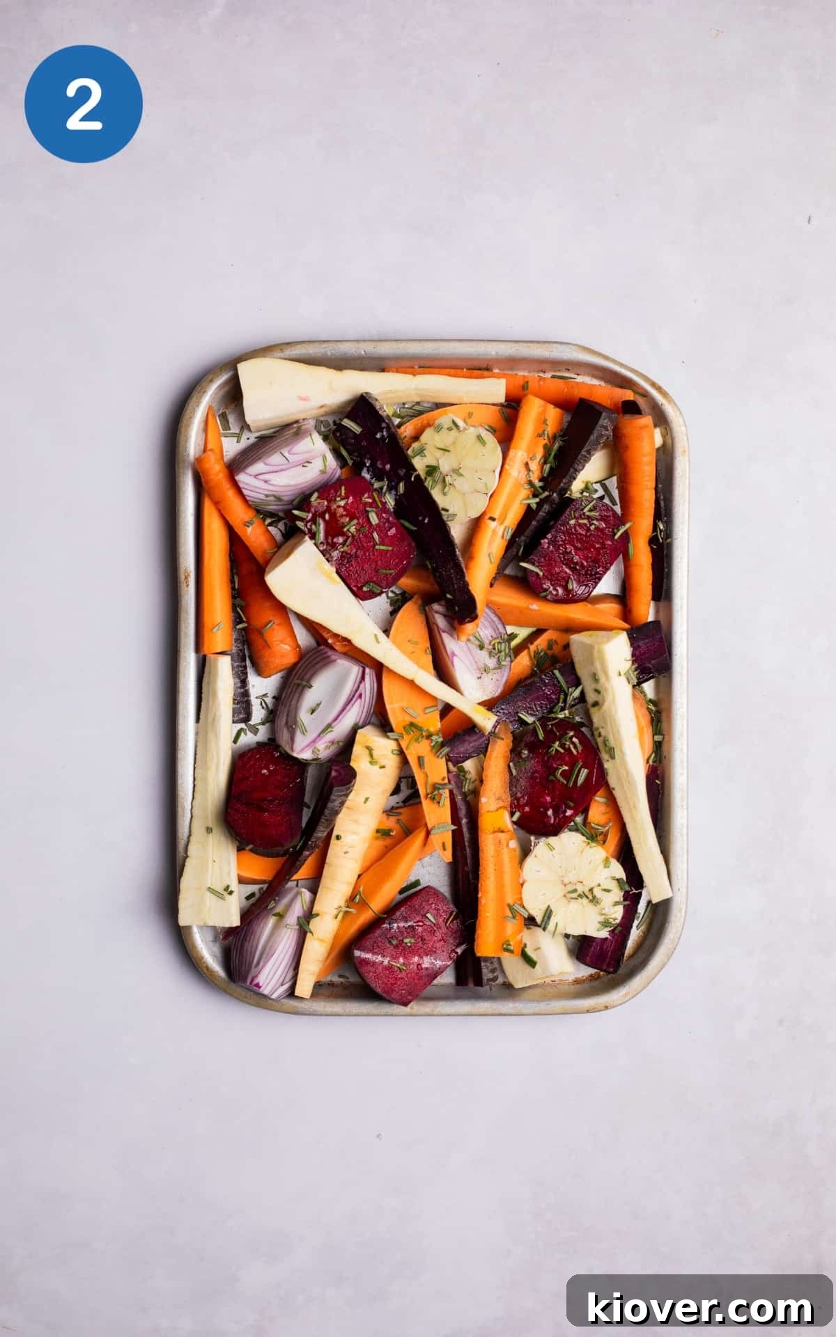 Chopped root vegetables tossed with olive oil and rosemary on a silver roasting tray.