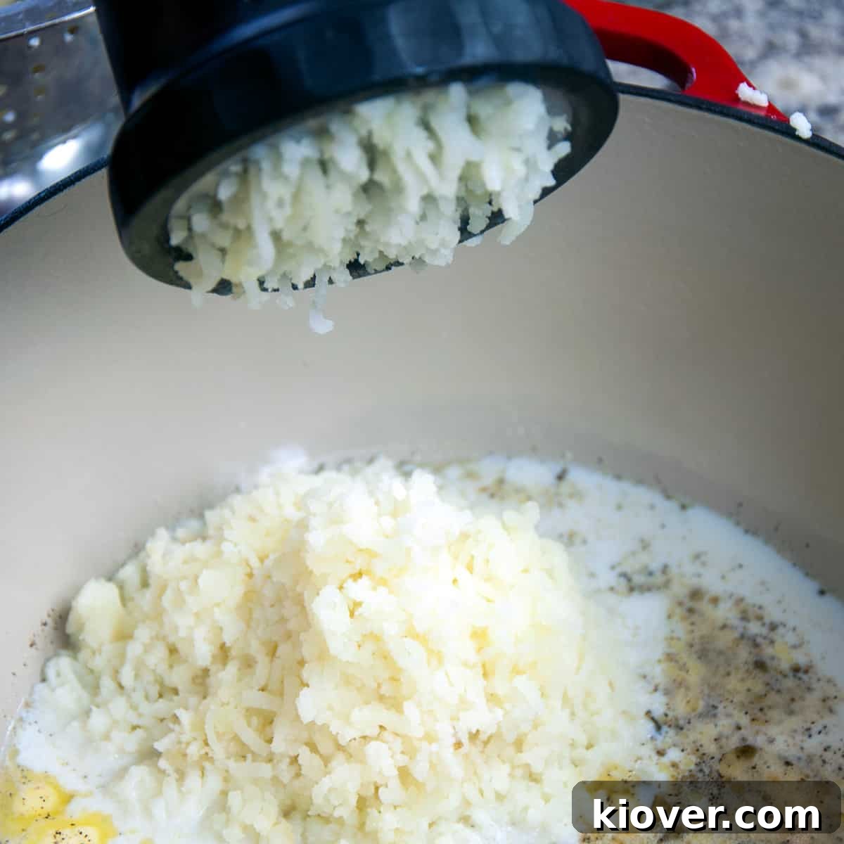 Cooked potatoes being pushed through a potato ricer into a bowl with melted butter and milk.