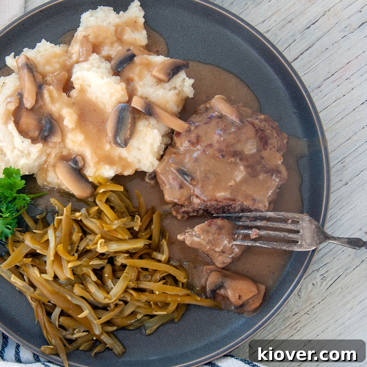 Finished Salisbury Steak Meal Salisbury Steak on a grey plate with green beans and mashed potatoes, garnished with fresh herbs.
