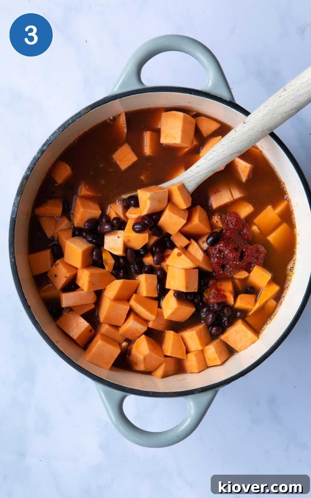 Hearty Sweet Potato and Black Bean Stew 6 A large pot on a white marble background, filled with pre-cooked ingredients for Sweet Potato and Black Bean Curry, including canned tomatoes, black beans, sweet potatoes, tomato puree, and vegetable stock, with a wooden spoon ready for stirring.