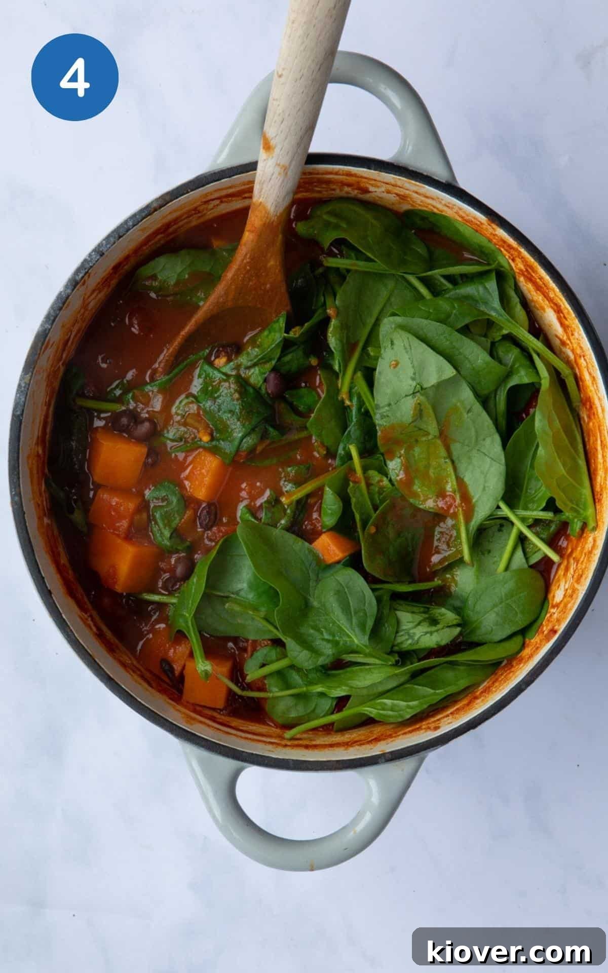 Hearty Sweet Potato and Black Bean Stew 7 Sweet Potato and Black Bean Curry after simmering for 25 minutes, presented in a large pot on a white marble background. Fresh spinach leaves are layered on top, ready to be stirred in and wilted before serving.