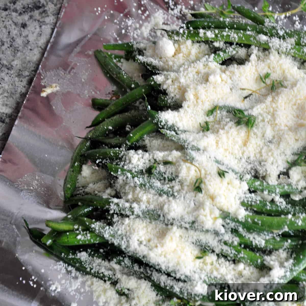 Grated Parmesan cheese generously sprinkled over green beans and thyme on a foil sheet.