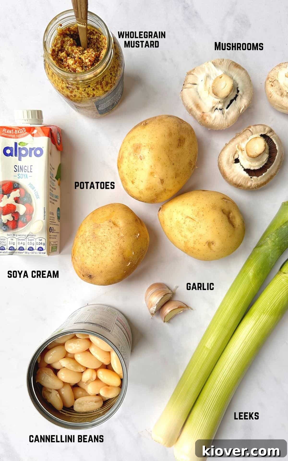 All the fresh ingredients for Vegan Mushroom Pie, including potatoes, mushrooms, leeks, garlic, wholegrain mustard, soya cream, and cannellini beans, artfully arranged on a white marble background.