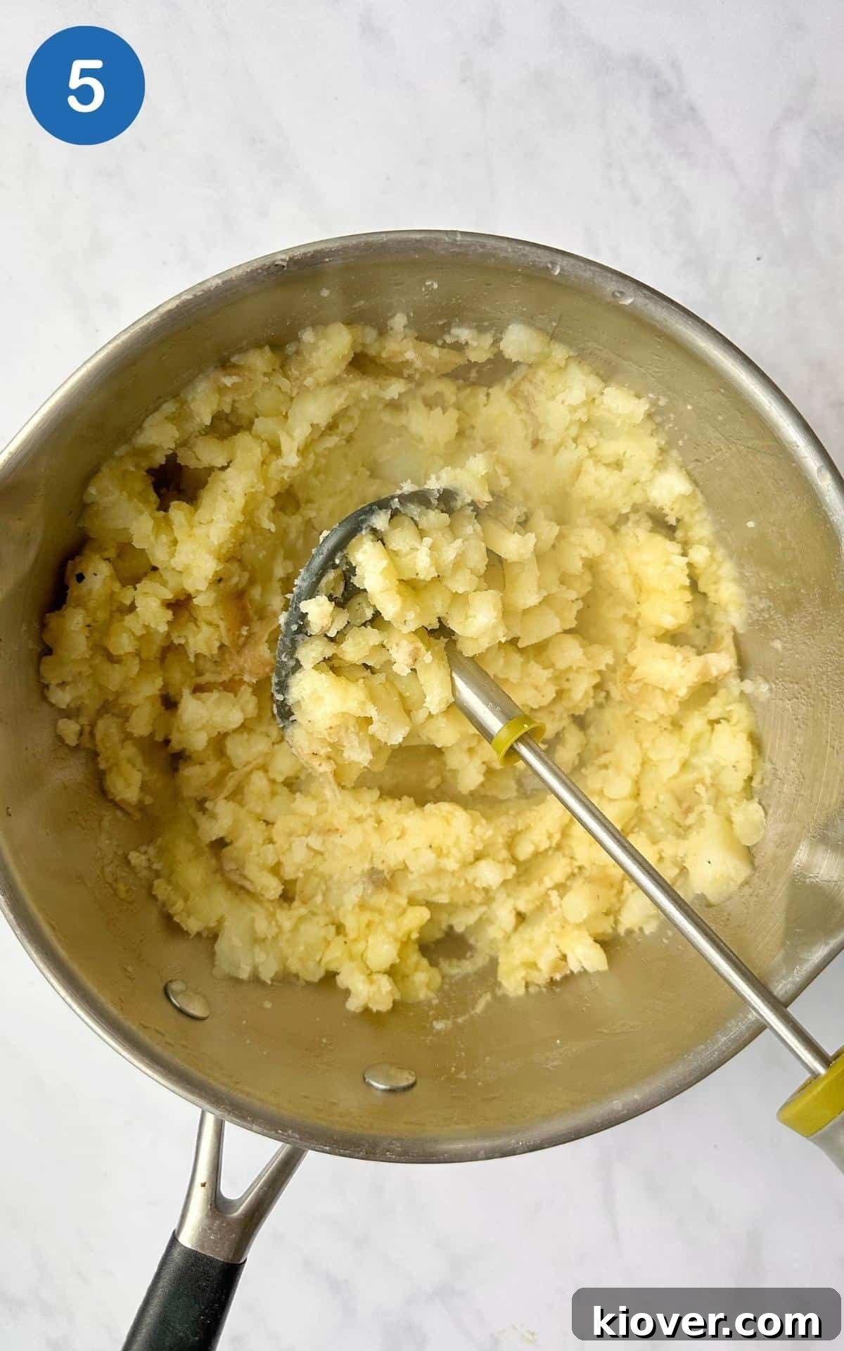 Perfectly mashed potatoes in a saucepan with a potato masher, having been enriched with olive oil, salt, and pepper, forming the soft, fluffy topping for the pie.