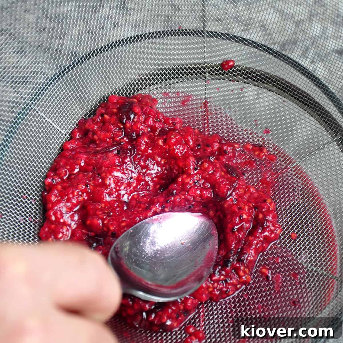 A fine mesh strainer with raspberries and black currant jelly being strained.