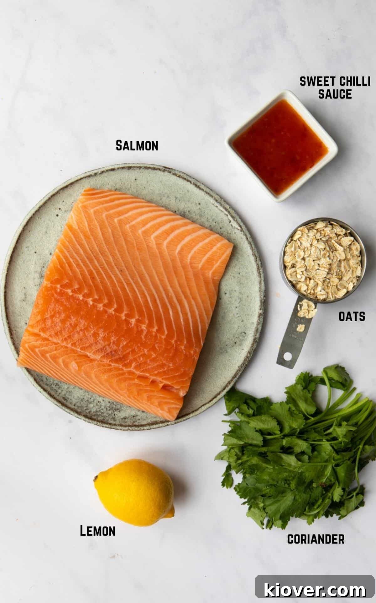 The five core ingredients for Sweet Chilli Salmon Fishcakes arranged artfully on a white marble background: fresh salmon fillets, vibrant lemon, a bottle of sweet chilli sauce, rolled oats, and a bunch of fresh coriander.