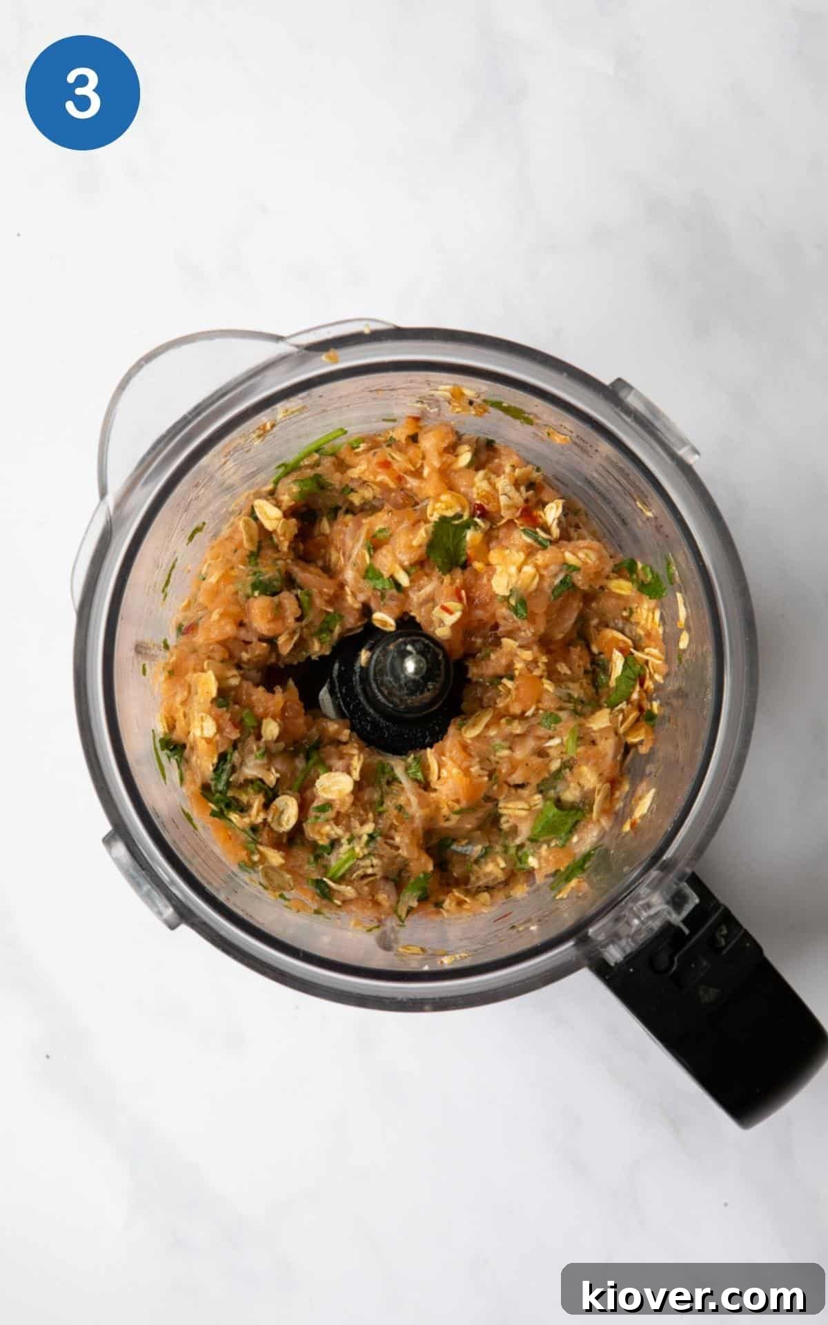 The perfectly blended mixture for Sweet Chilli Salmon Fishcakes, a vibrant salmon-colored paste with visible flecks of green coriander, resting inside a food processor bowl on a sleek white marble background.