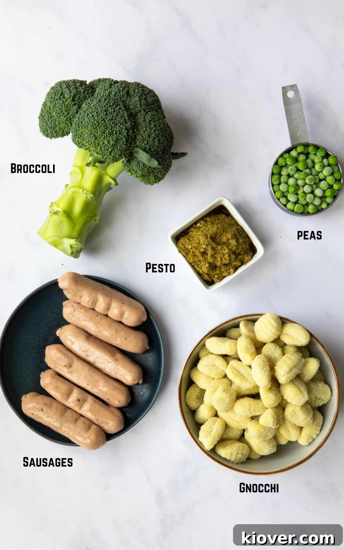 Savory Sausage Pesto Gnocchi 3 Image showing all the ingredients needed to make Sausage Pesto Gnocchi laid out on a white marble background, including gnocchi, sausages, broccoli, pesto, and peas.