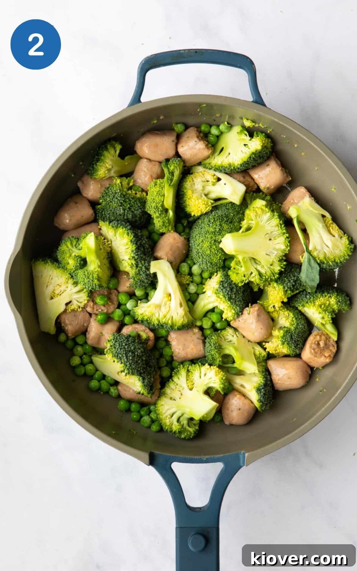 Savory Sausage Pesto Gnocchi 5 Raw broccoli, sausage and peas in a large non stick frying pan before being cooked. Pan is positioned on a white marble background.