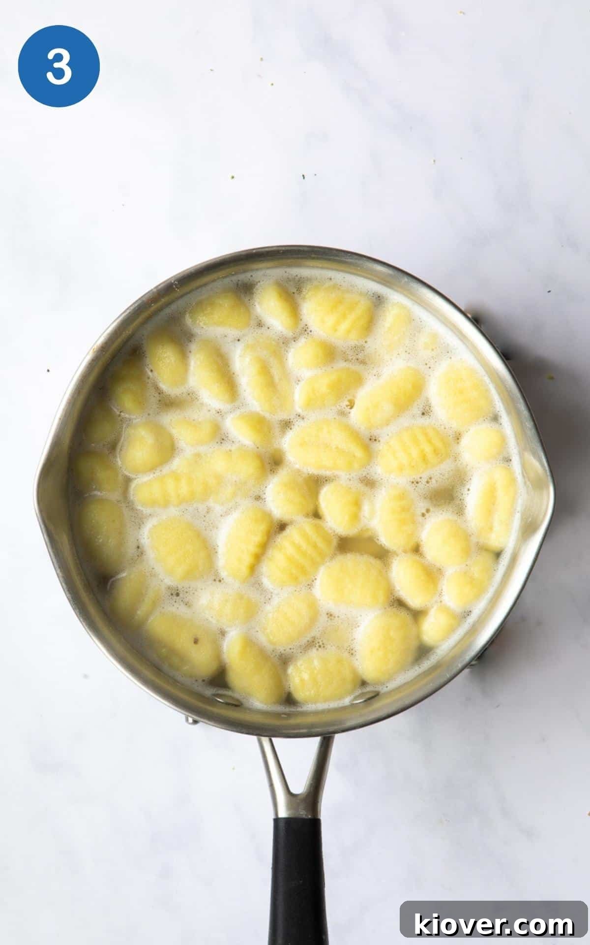 Savory Sausage Pesto Gnocchi 6 Gnocchi being boiled in salted water in a saucepan. Displayed on a white marble background.
