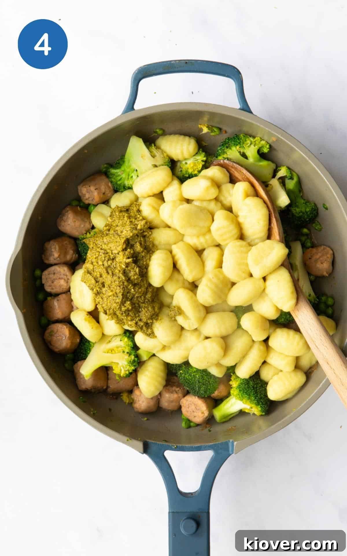 Savory Sausage Pesto Gnocchi 7 Gnocchi and pesto being added to a large non stick frying pan. There is also a wooden spoon in the image to help combine the ingredients. Pan is displayed on a white marble background.