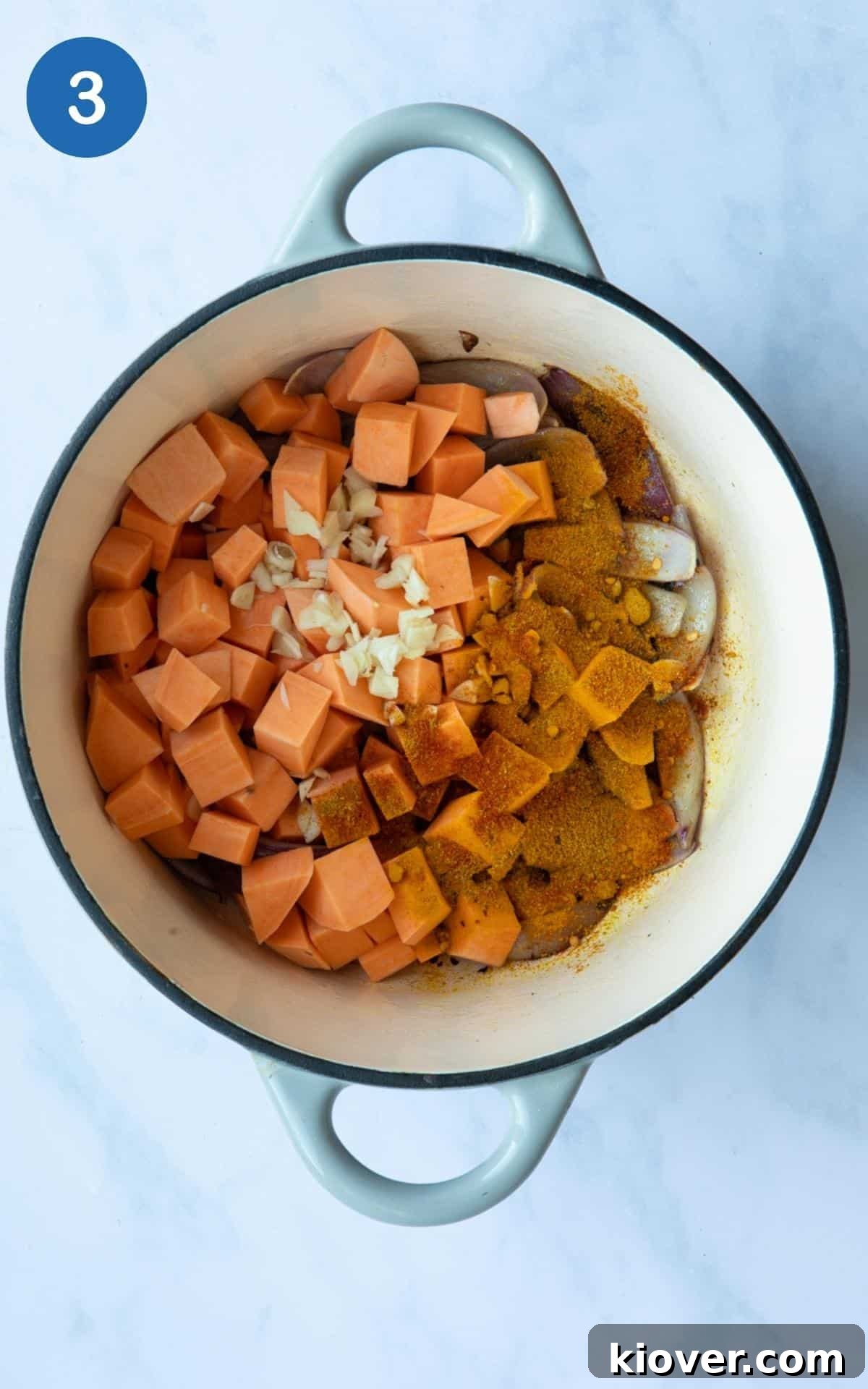 Uncooked chopped sweet potato, onions, and garlic inside a large oven-proof pot, displayed on a white marble background.