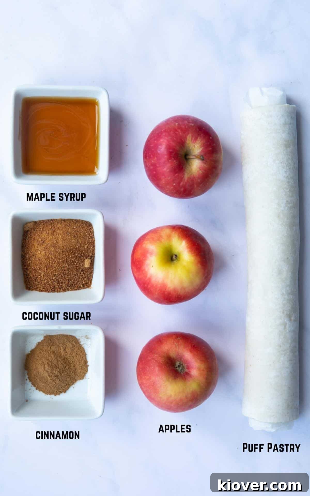 Five core ingredients - apples, maple syrup, ground cinnamon, coconut sugar, and a sheet of puff pastry - are artfully arranged on a white marble background, showcasing their natural beauty and simplicity.