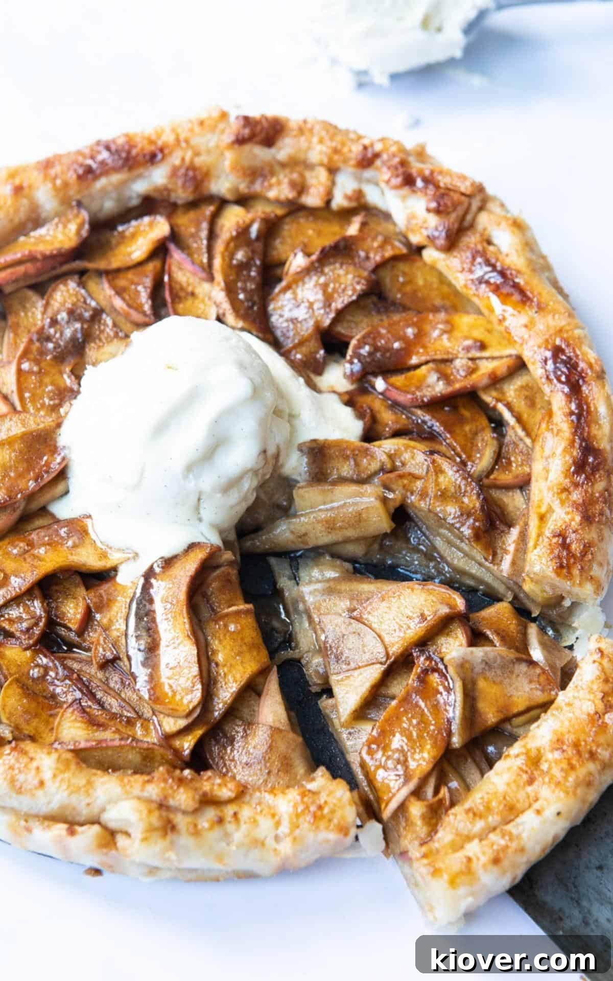 A perfectly baked slice of Puff Pastry Apple Tart is being gently cut from the main tart, which features a melting scoop of vanilla ice cream in its center. The delicious dessert is presented on a clean white marble background.