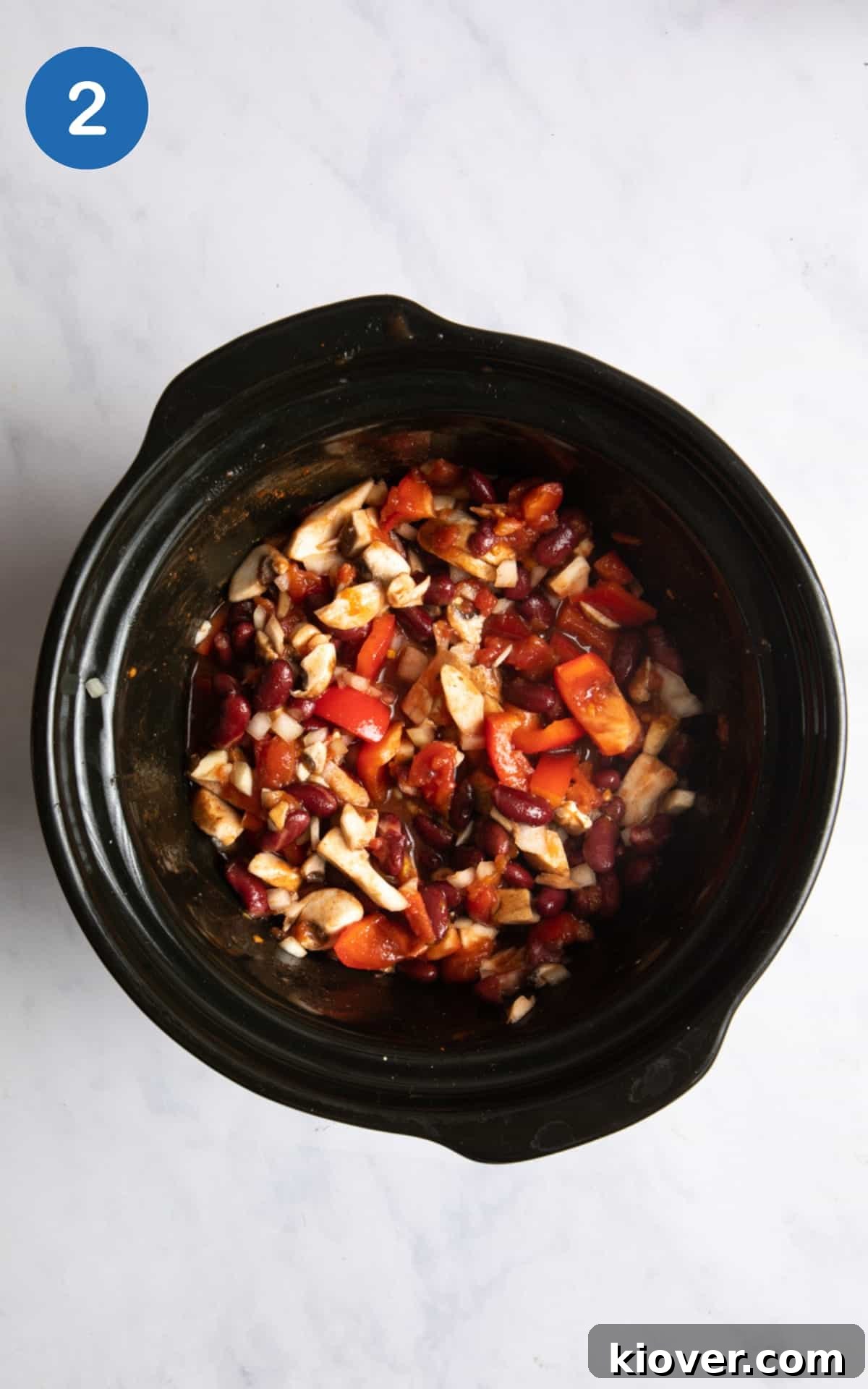 An overhead shot of all the vegan chilli ingredients—chopped vegetables, beans, tomatoes, and spices—neatly layered inside a slow cooker, ready for cooking.