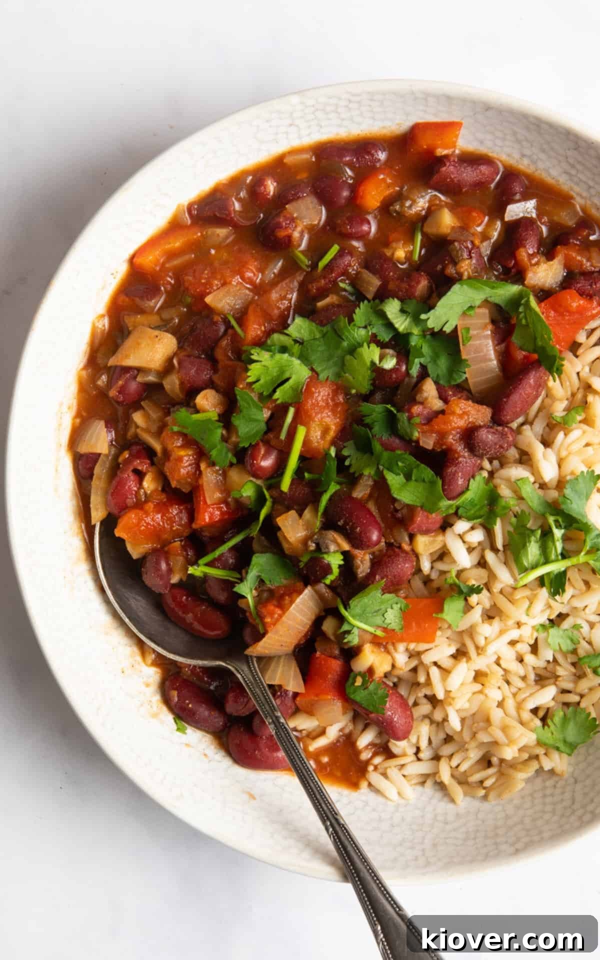 A serving of rich slow cooker vegan chilli in a white bowl, garnished with fresh coriander, accompanied by a side of fluffy rice.