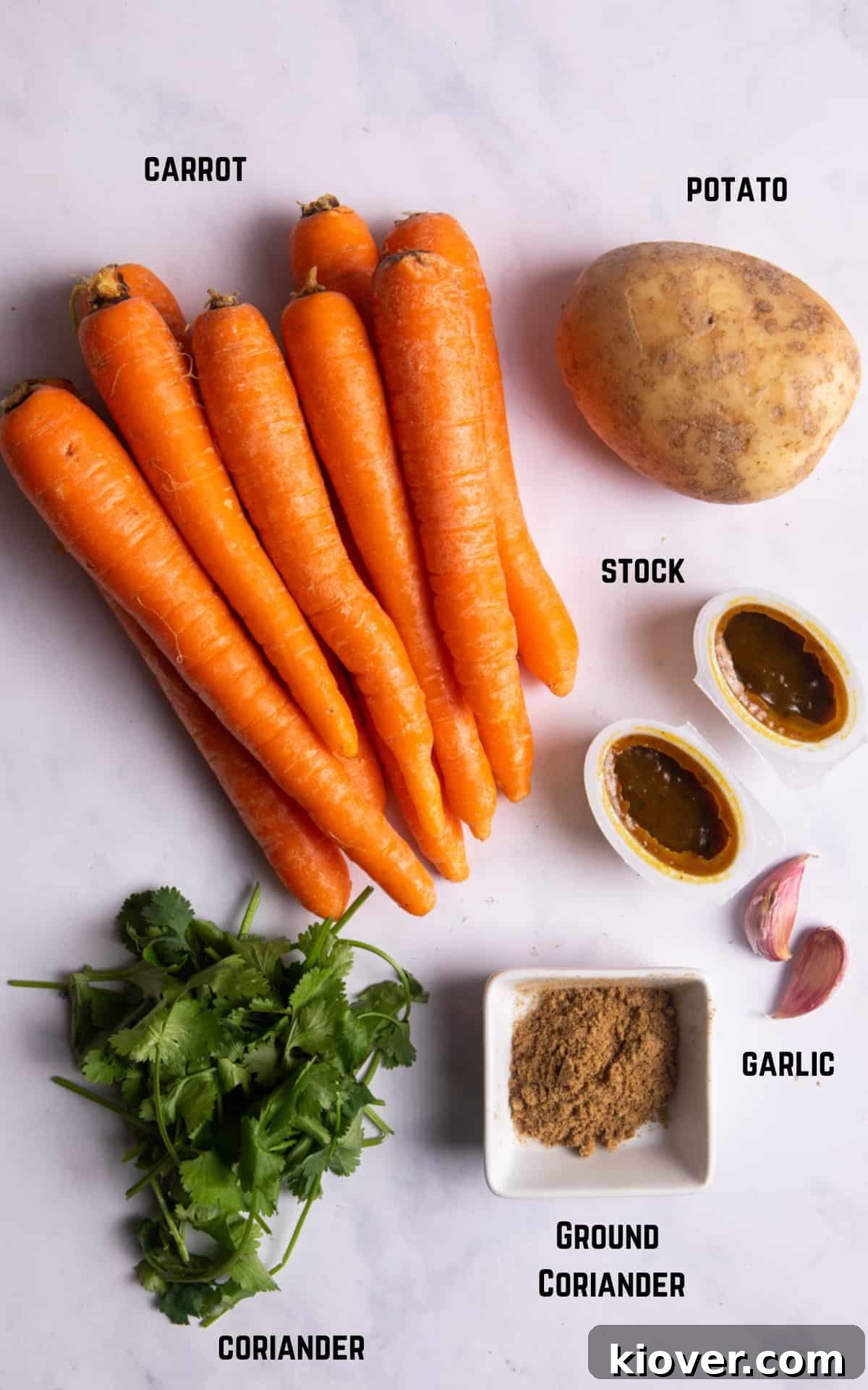 Fresh ingredients laid out on a white marble background, including carrots, potato, garlic, vegetable stock, ground coriander, and fresh coriander, ready to make the soup.