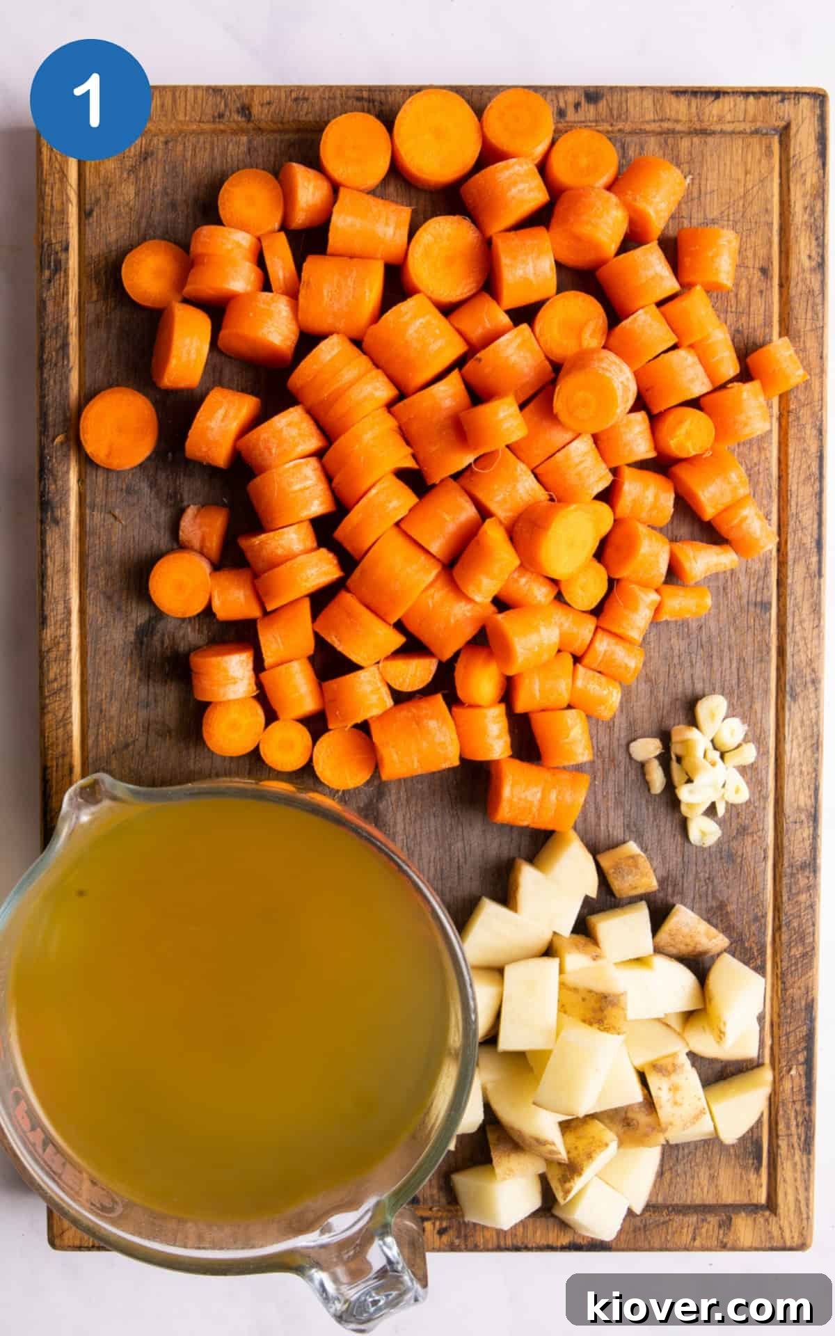 Roughly chopped carrots, potato, and garlic are arranged on a dark wooden chopping board. A measuring jug filled with vegetable stock is next to them, all set on a white background.