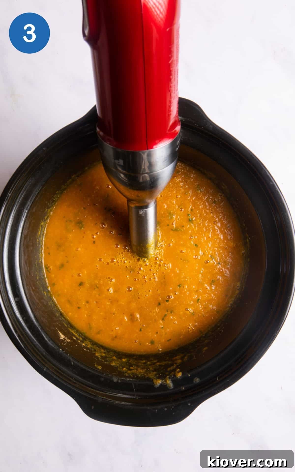 Soup ingredients are being blended directly in the slow cooker using a hand blender after being cooked. The slow cooker is on a white background.