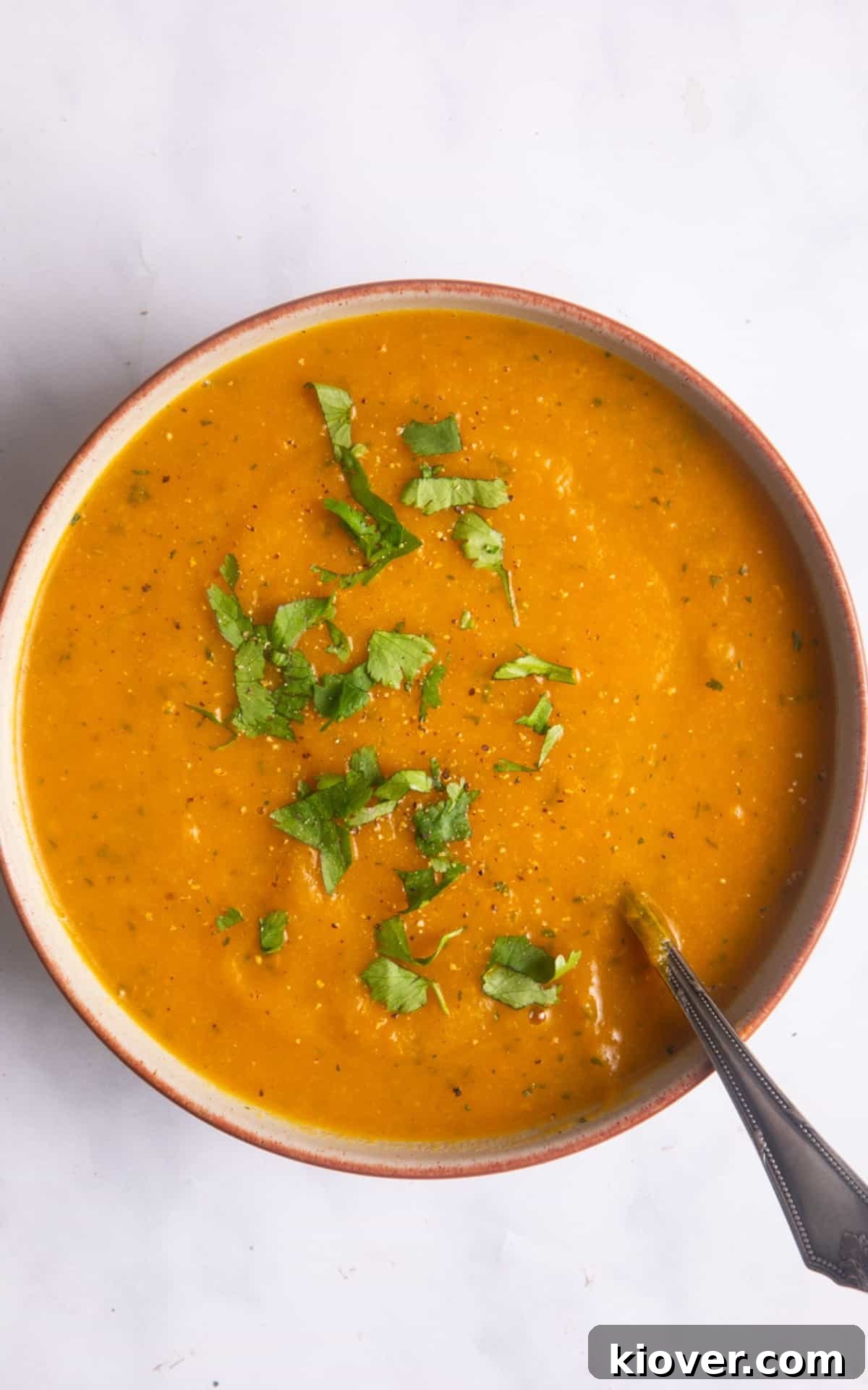 Overhead shot of a vibrant Carrot and Coriander Soup served in a white bowl with an orange rim, garnished with fresh coriander and a silver spoon, presented on a white marble background.
