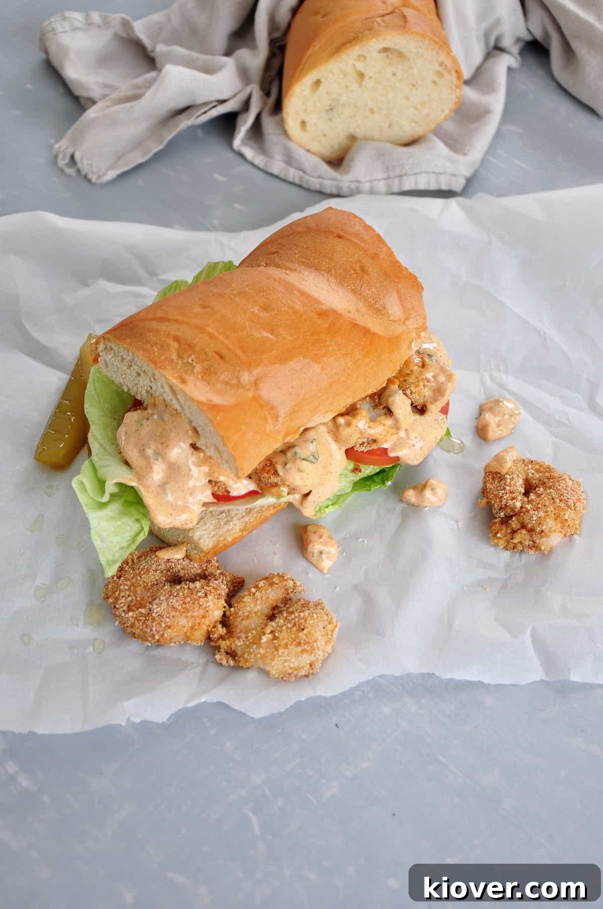 Shrimp Po' Boy A beautifully presented, finished Baked Shrimp Po' Boy sandwich, generously topped with remoulade sauce, with a traditional French bread loaf wrapped in cloth in the soft background.
