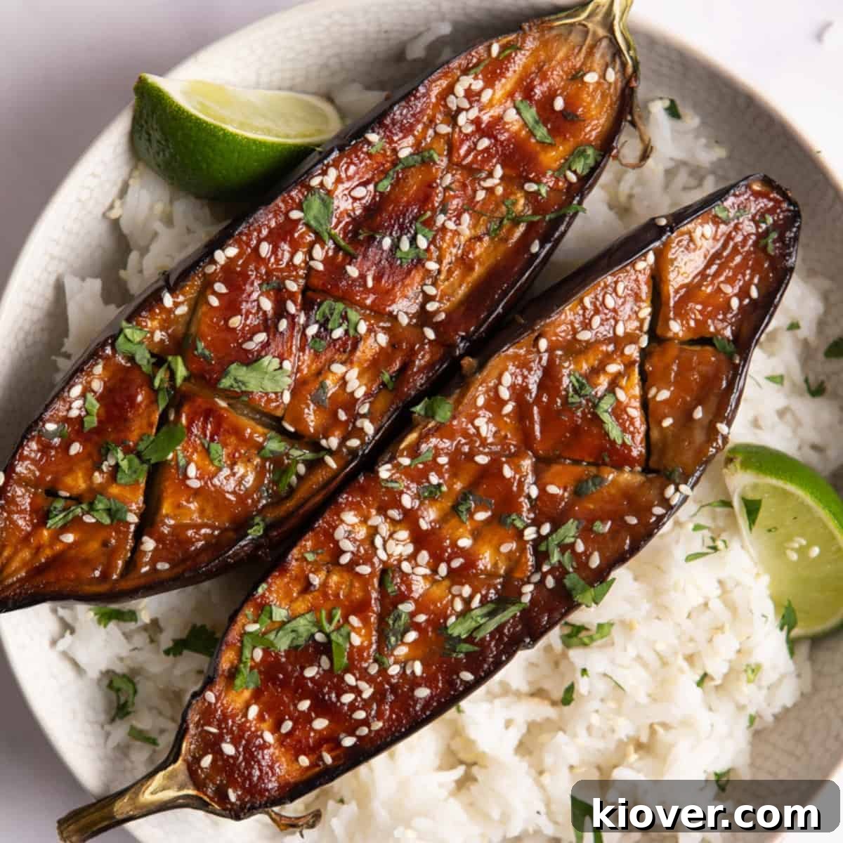 Perfectly Sweet and Savory Miso Glazed Aubergine Nasu Dengaku 2 Two beautifully miso-glazed aubergine halves, glistening with their savory-sweet coating, served over a bed of fluffy white rice. Fresh lime wedges are artfully placed beside the aubergines, suggesting a hint of citrus. The white bowl rests on a clean, white surface, highlighting the vibrant colors of the dish.