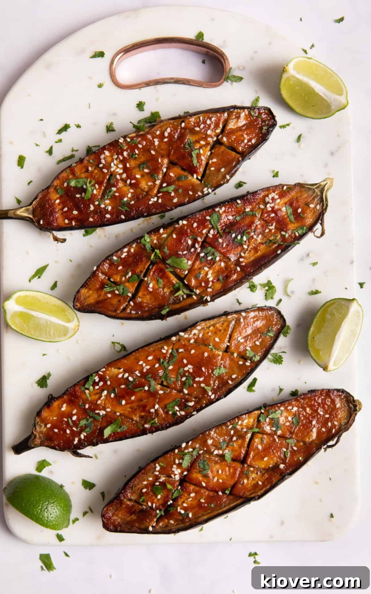 Perfectly Sweet and Savory Miso Glazed Aubergine Nasu Dengaku 8 Two beautifully plated miso-glazed aubergine halves, generously topped with toasted sesame seeds, vibrant fresh coriander, and bright lime wedges. The dish is presented on a rustic white chopping board, emphasizing its freshness and appealing garnishes.