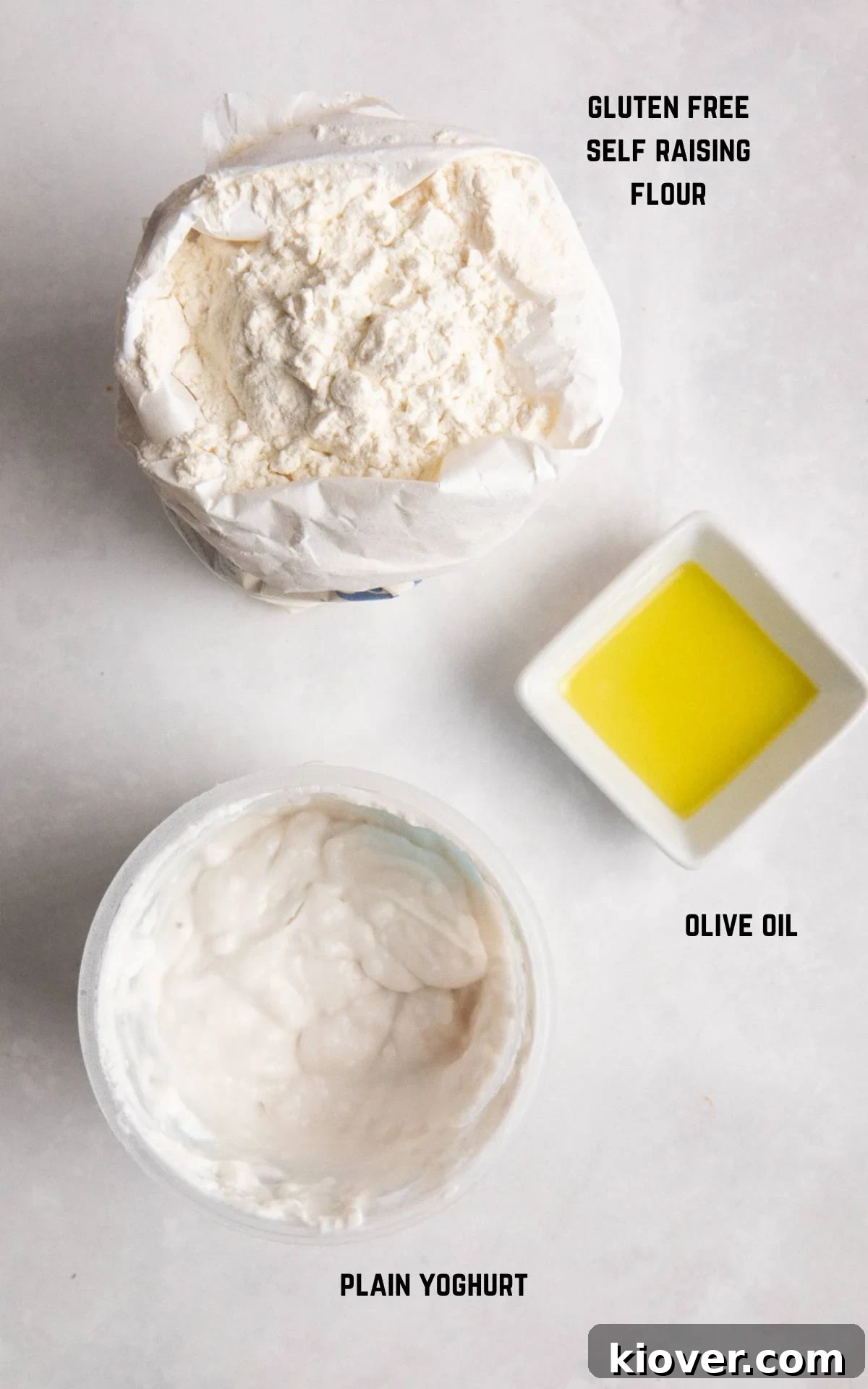 Essential ingredients for gluten-free flatbread: a bowl of flour, a container of plain yogurt, and a bottle of olive oil, neatly arranged on a white marble surface.